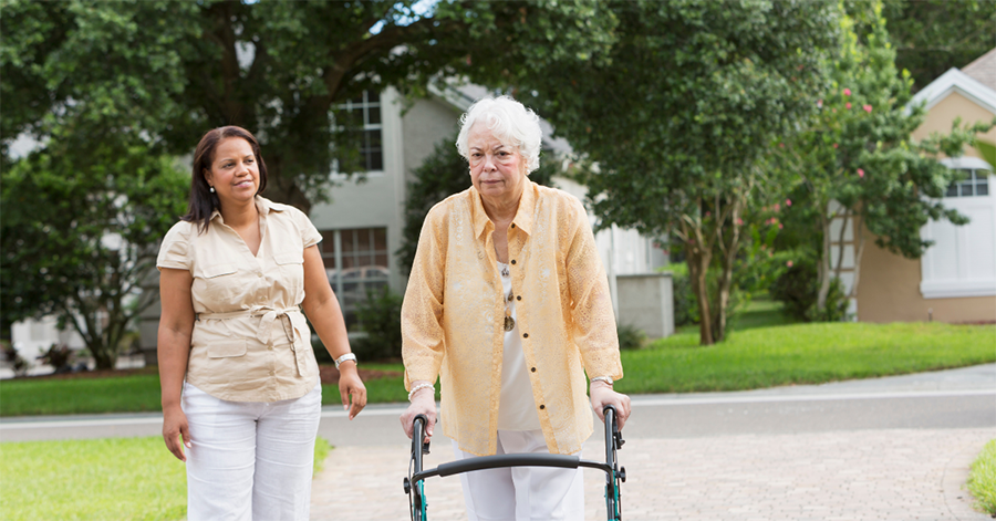 A woman on a walk with her mother, who uses a walker, representing an adult child proactively caring for an aging parent.