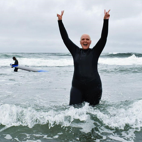 A person participating in the First Descents program, stands in the ocean. The person is wearing a black wet suit and giving the peace sign.