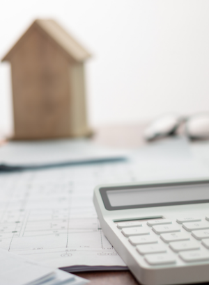 A calculator sits on a stack of papers with a tiny wooden home in the background, representing cancer finance and estate planning