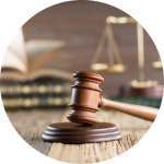 A wooden gavel sits on a table with law books and the scales of justice faded in the background