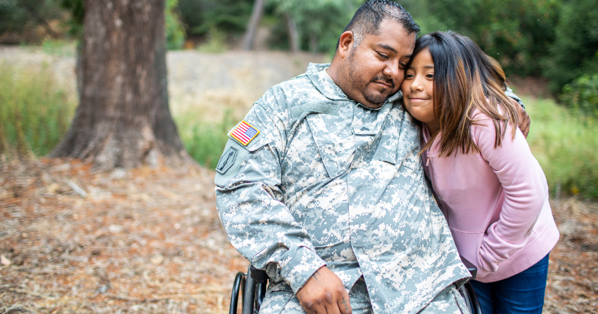 A veteran sitting in a wheelchair hugs his daughter. They are outside.