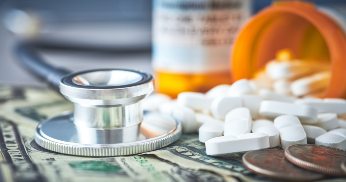 A pill bottle, filled with white pills, is spilling over onto a pile of money with a stethoscope on top of it.