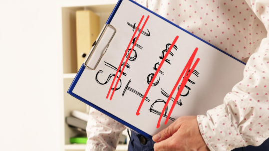 A person wearing a white shirt with pink dots is carrying a clip board with a piece of paper that says "short term plan" and the words are crossed out in red.