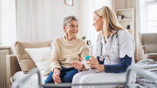 A home health caregiver in scrubs sits beside an older woman on a couch, holding a pill container and speaking with her in a bright, comfortable living room.
