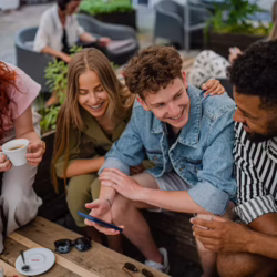 Four young adults sit together outdoors at a café or patio table, smiling and looking at a smartphone. They appear engaged in a friendly conversation while enjoying drinks in a relaxed social setting.